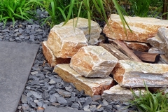 Fireplace from Limestone in Designed Alpine Backyard Garden with Tiled Footpath. Stone Fire Pit from Stone with Firewood near Garden Path from Tiles and Gravel. Landscaped Backyard Design.